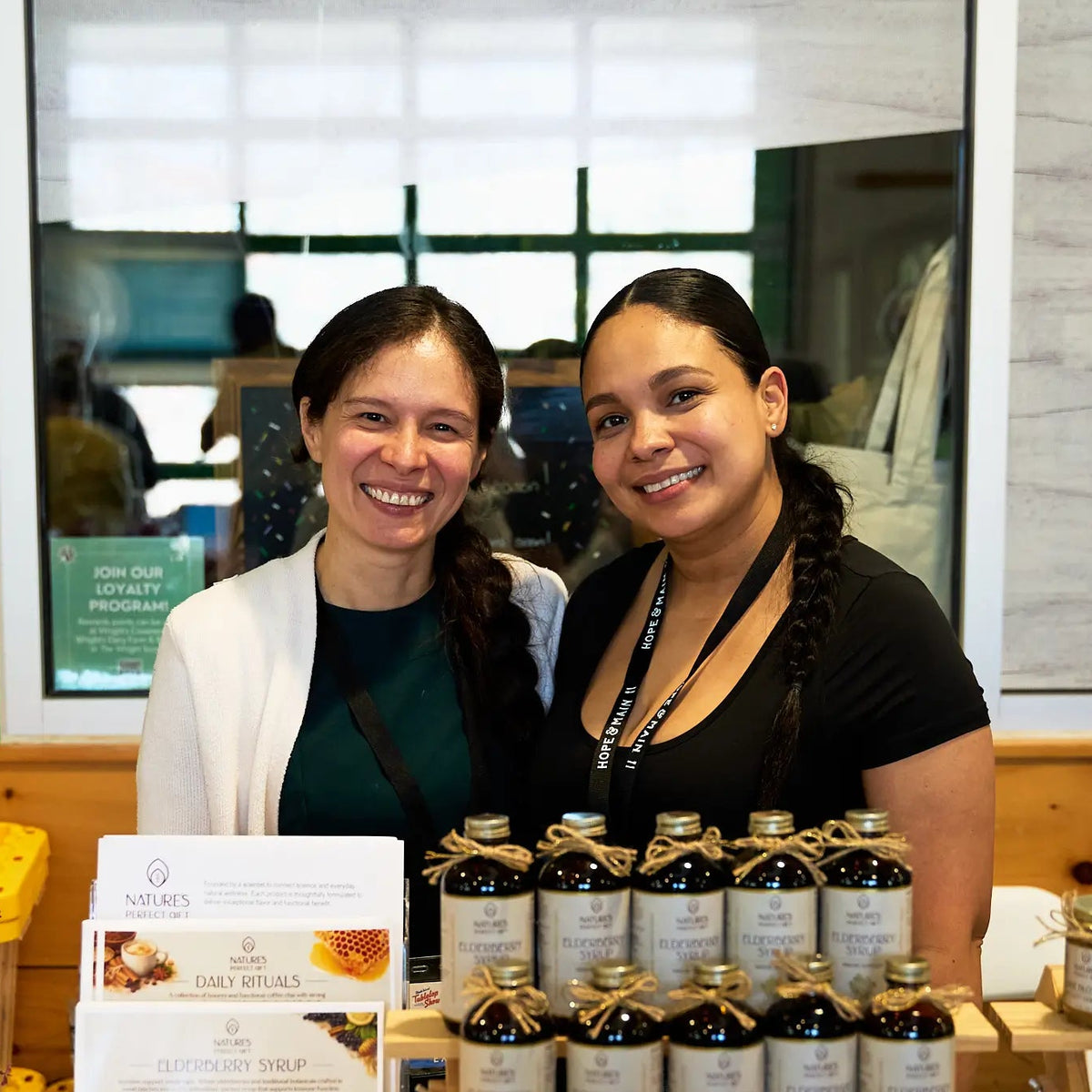 Two smiling women Dr. Borgas and CEO standing behind a display of Nature's Harmony brand syrup bottles. The bottles are dark glass with white labels and gold caps, tied with twine, and arranged on a table with a branded card holder in front.