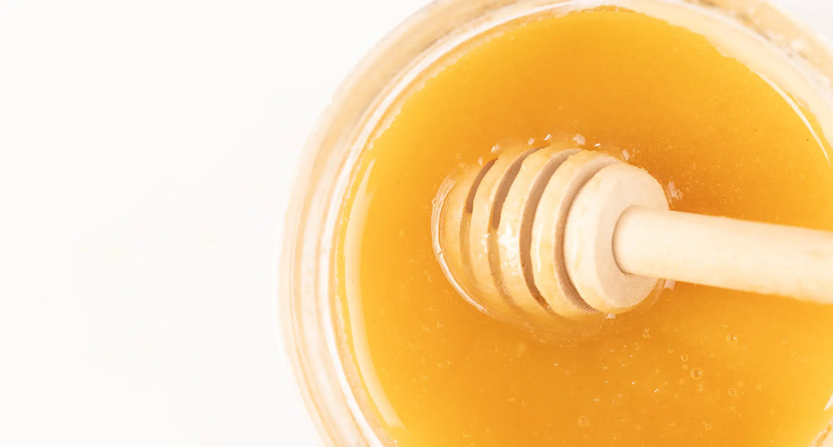 Top-down close-up of golden honey in a clear glass jar with a wooden honey dipper on a white background.