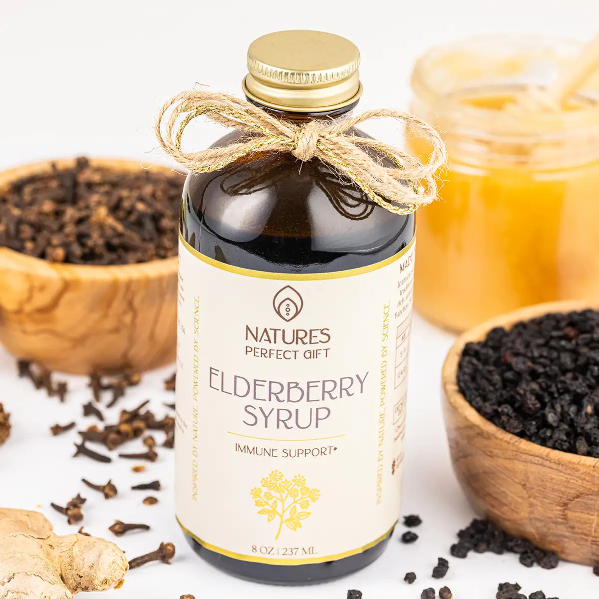 A bottle of Nature's Perfect Gift Elderberry Syrup with a gold cap and twine bow, surrounded by bowls of ingredients including black elderberries, ginger, and honey on a white background.
