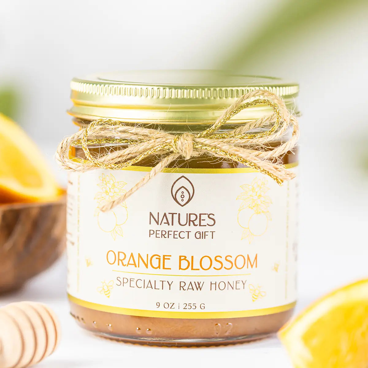 A glass jar of Natures Perfect Gift Orange Blossom Specialty Raw Honey, featuring a gold metal lid, a gold and white label, and a rustic twine bow. The jar is styled with orange slices and a wooden honey dipper in the background.
